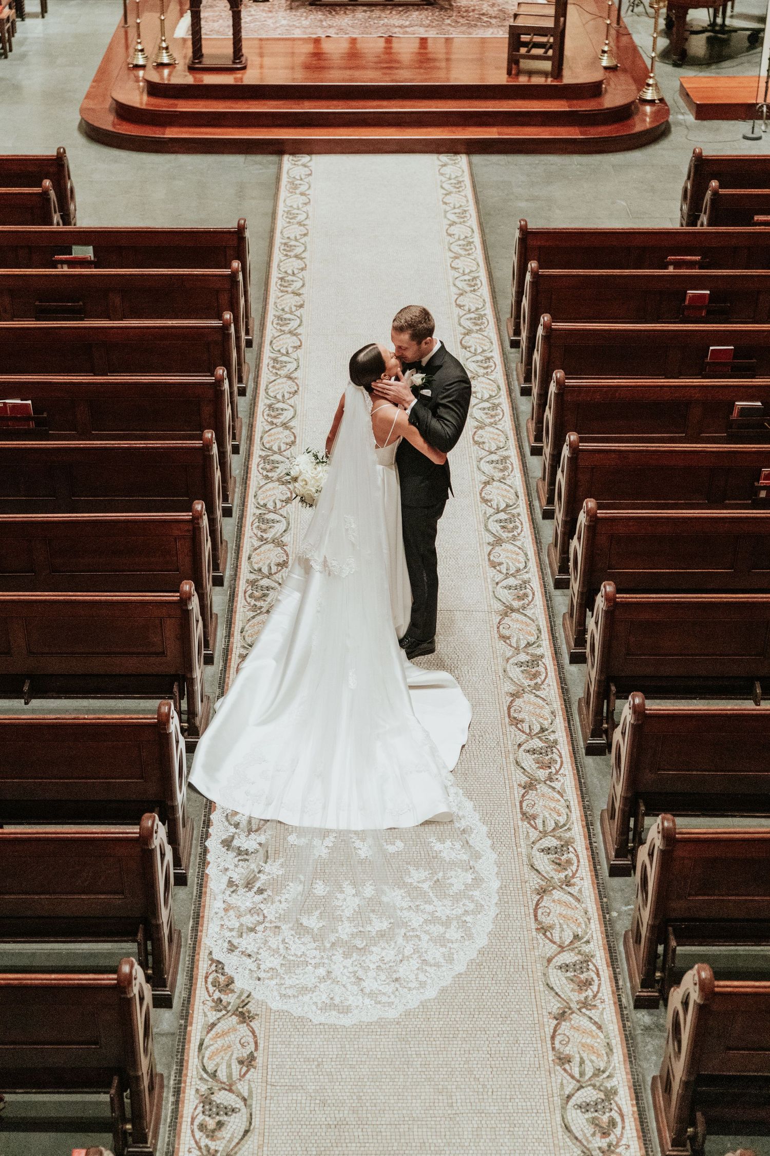 Wedding couple shares an intimate moment from above in a church aisle with wooden pews and ornate carpeting.