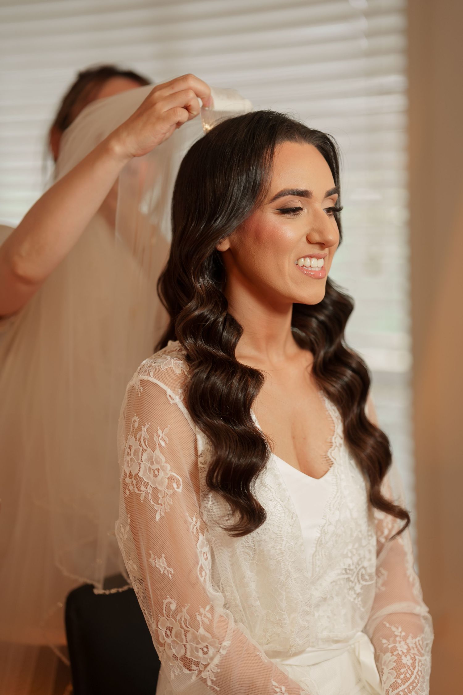 A bride in a white lace dress with long sleeves laughs joyfully while preparing for her wedding day.