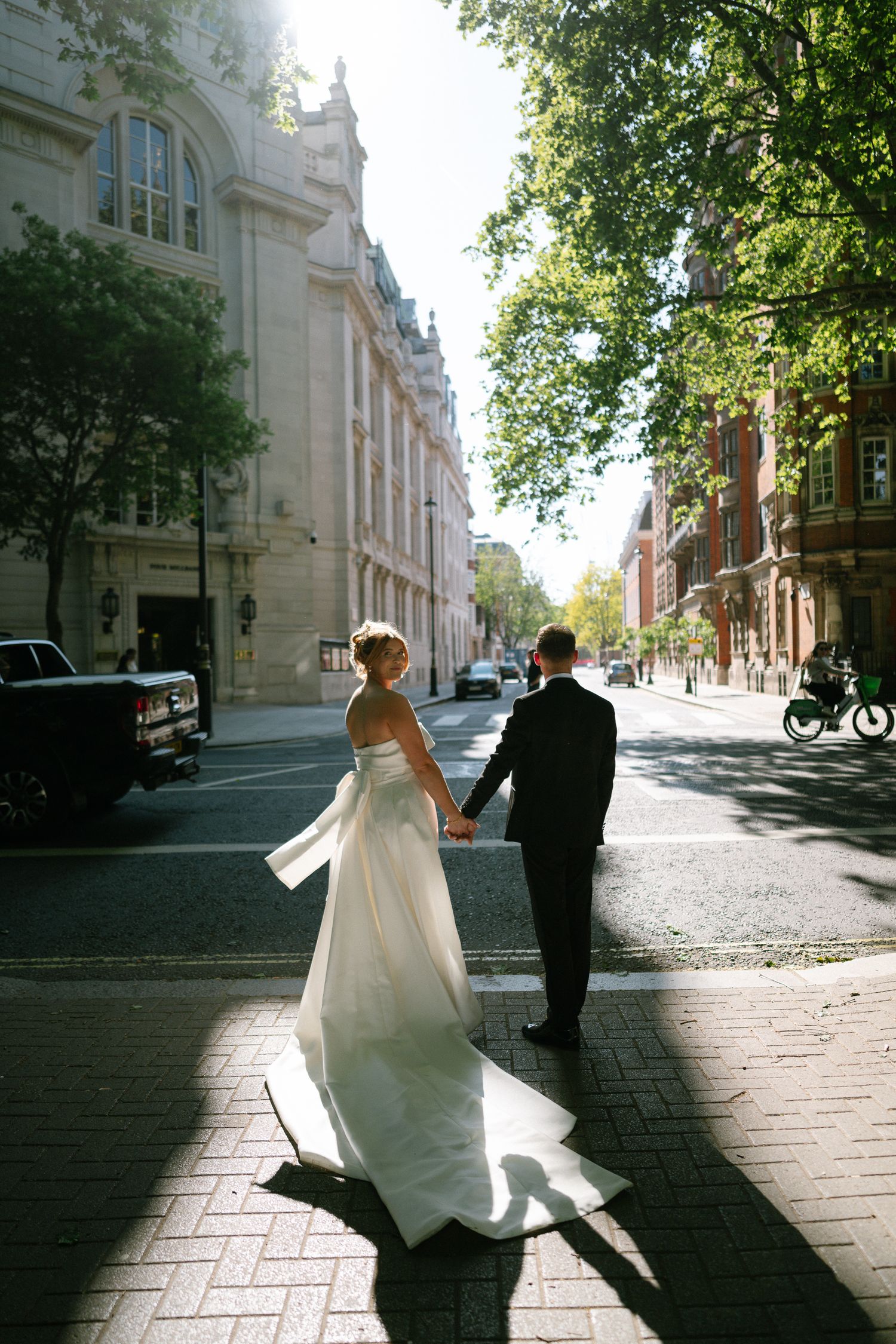 A couple in wedding attire runs down a city street holding hands with a classical brick building facade in the background.