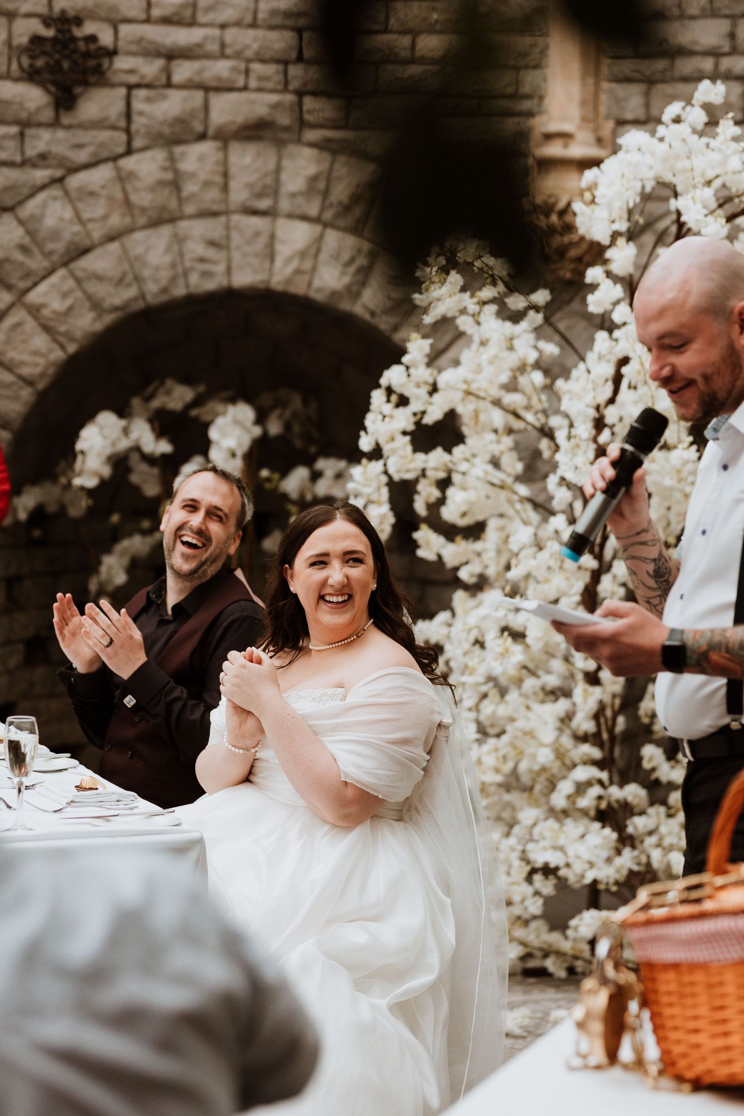 Wedding guests laugh and smile during a joyful moment surrounded by white floral decorations and stone walls at Tortworth Court