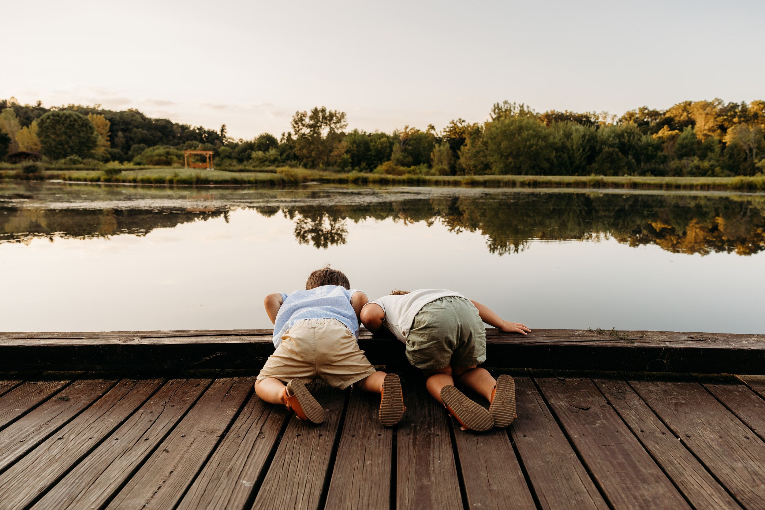 Two figures lie on their stomachs on a wooden dock overlooking a calm lake with autumn trees reflecting in the water.