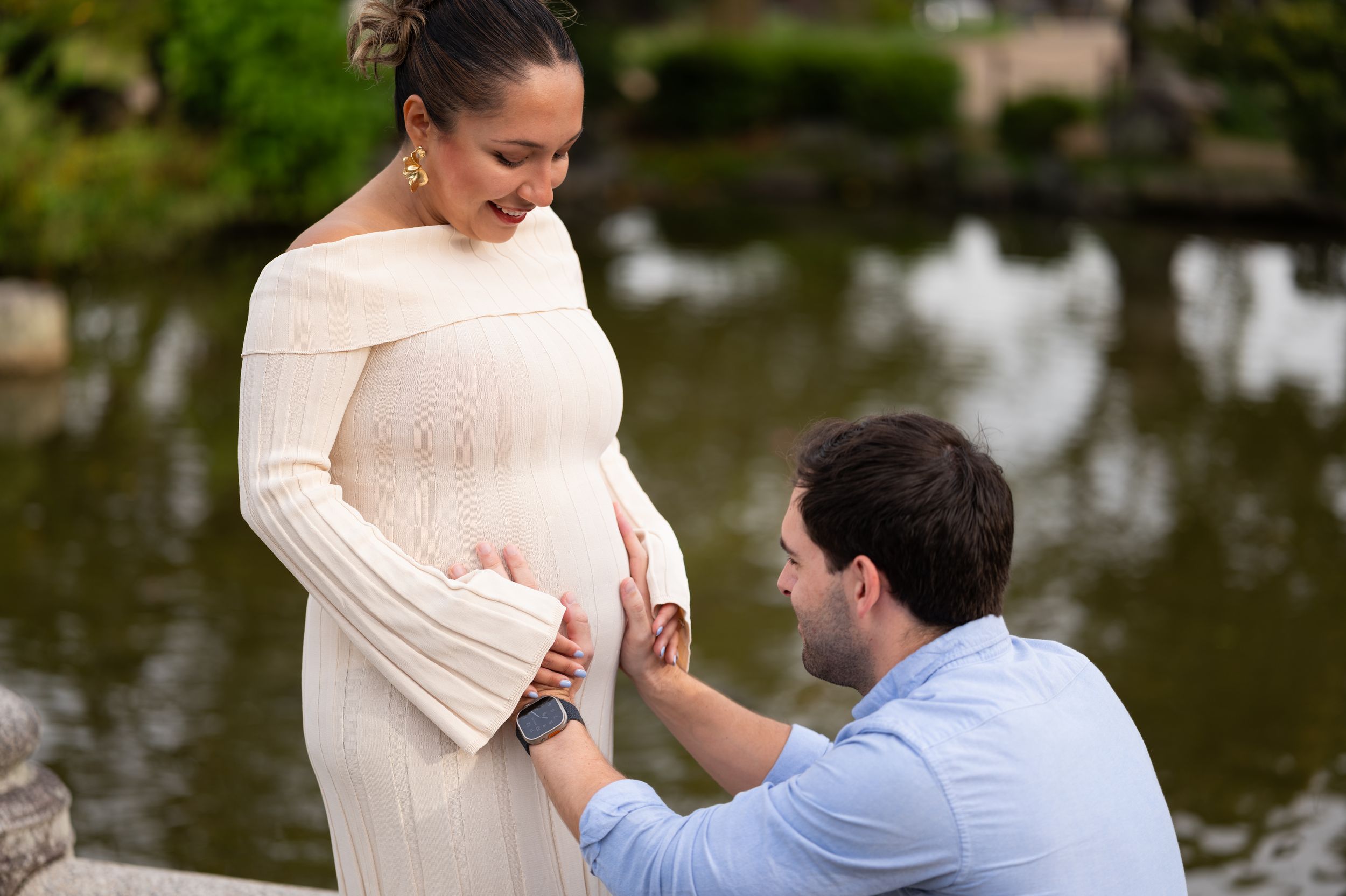 Man kneeling to touch pregnant woman's belly by a reflective pond in a Japanese garden.