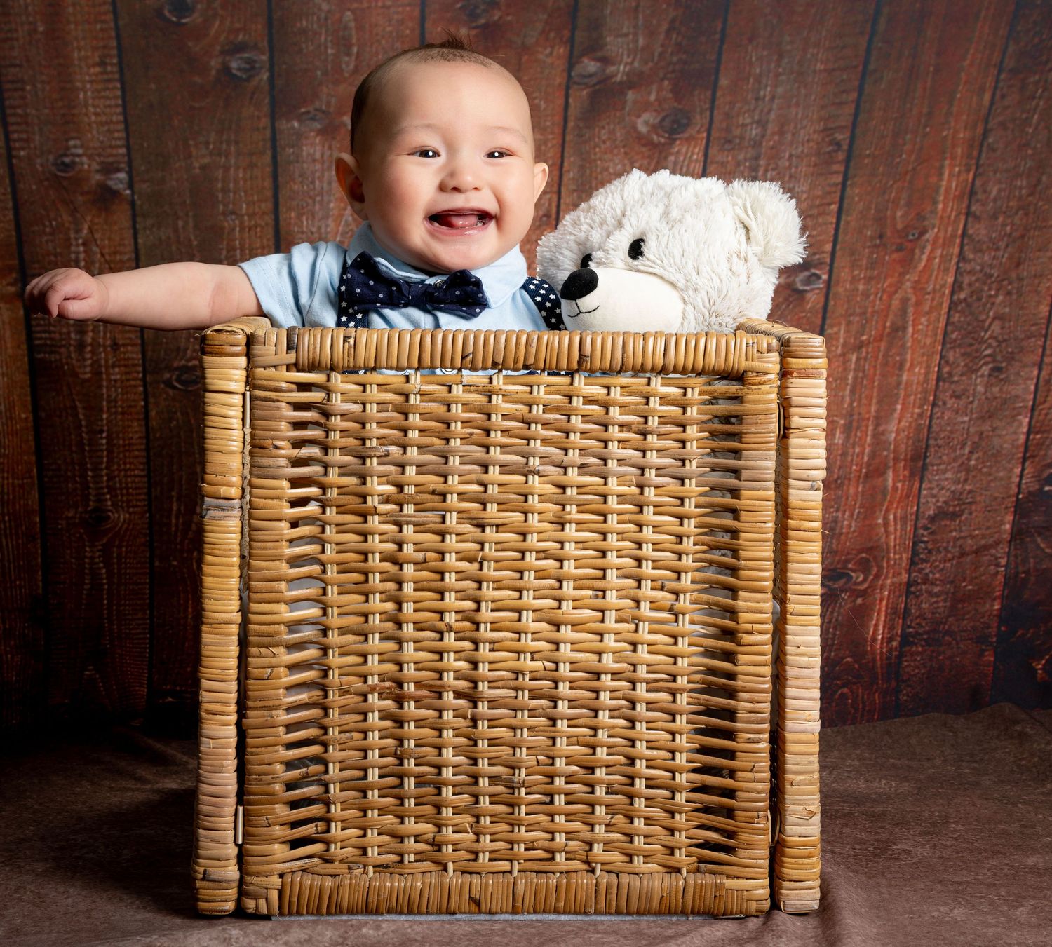 A happy baby sits in a woven wicker basket against a rustic wooden background with a white teddy bear.