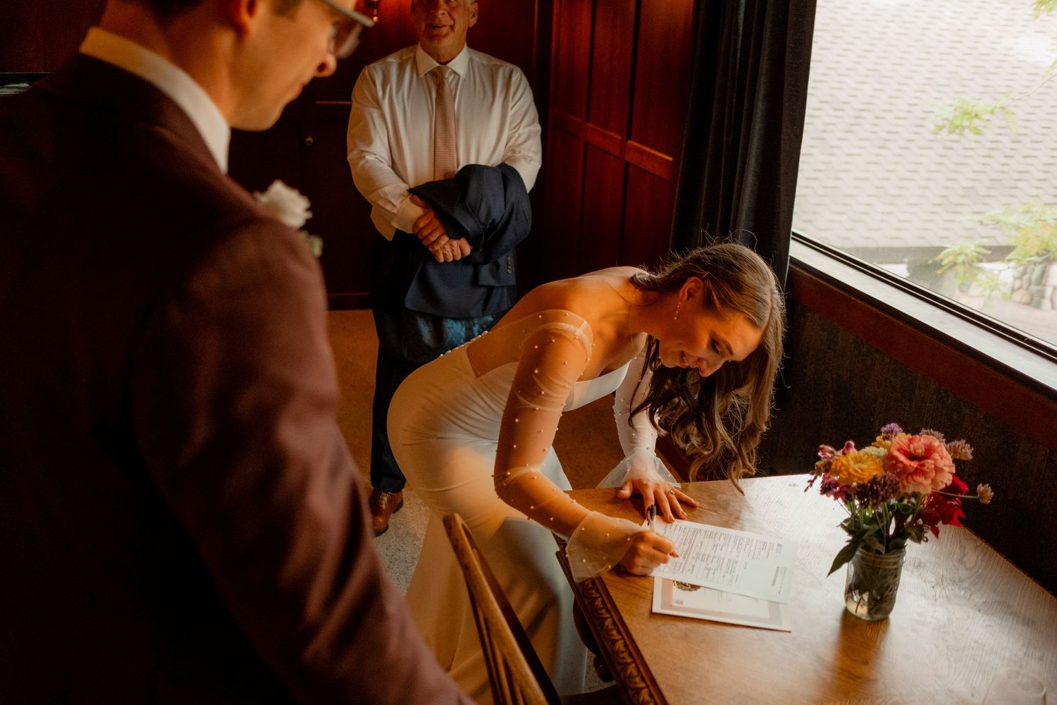 Couple signing documents together at a wooden table while witnesses observe at an intimate wedding ceremony.