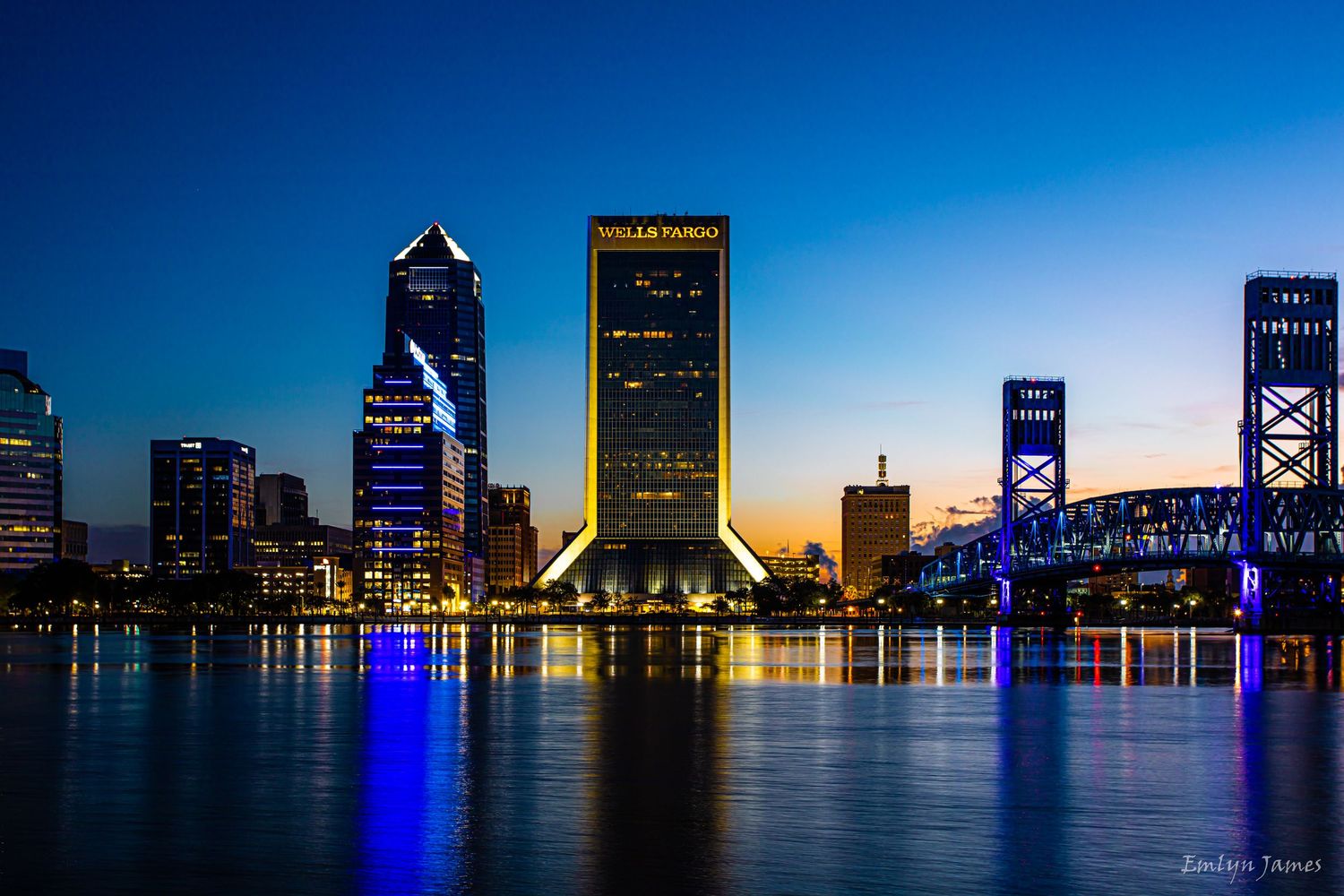 Jacksonville Florida skyline at dusk reflecting in St Johns River with illuminated Main Street Bridge and buildings.