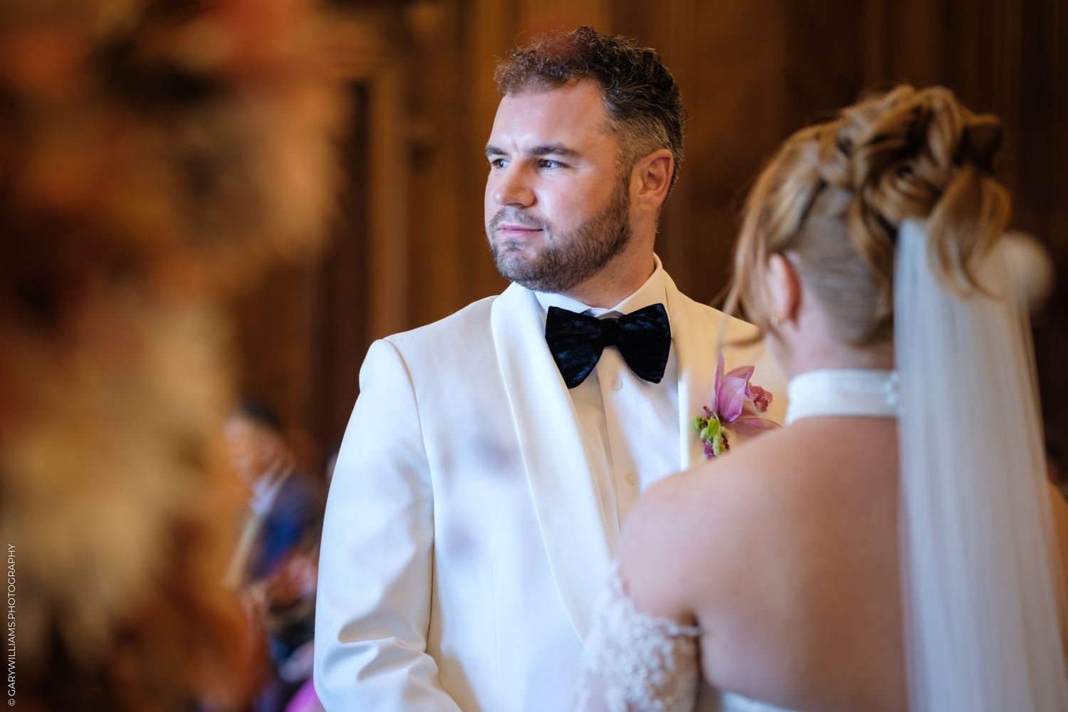 Groom in white tuxedo with black bowtie and boutonniere smiles during wedding ceremony.