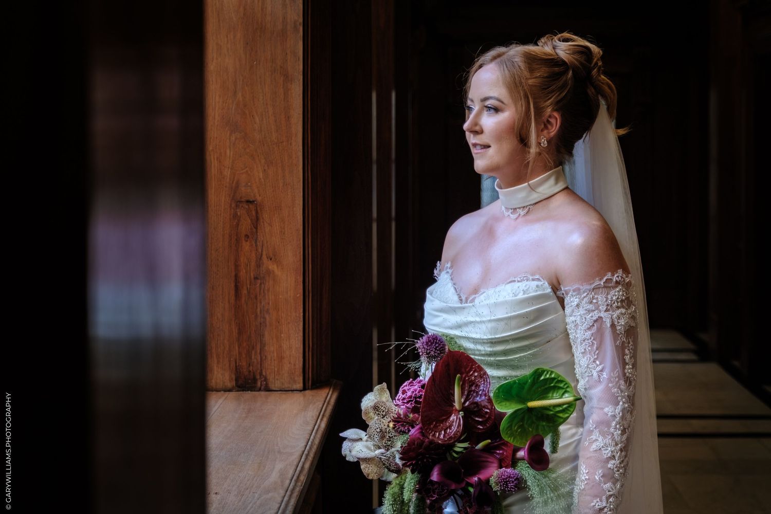 A bride in a white gown with lace sleeves poses near a wooden window frame in dramatic lighting.