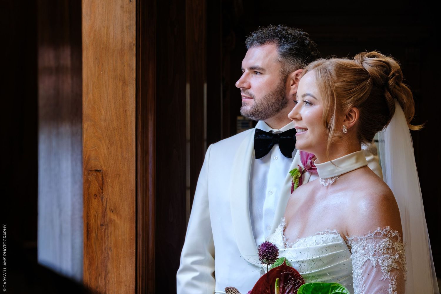 A bride in a white off-shoulder gown and groom in white tuxedo stand together during their wedding ceremony.