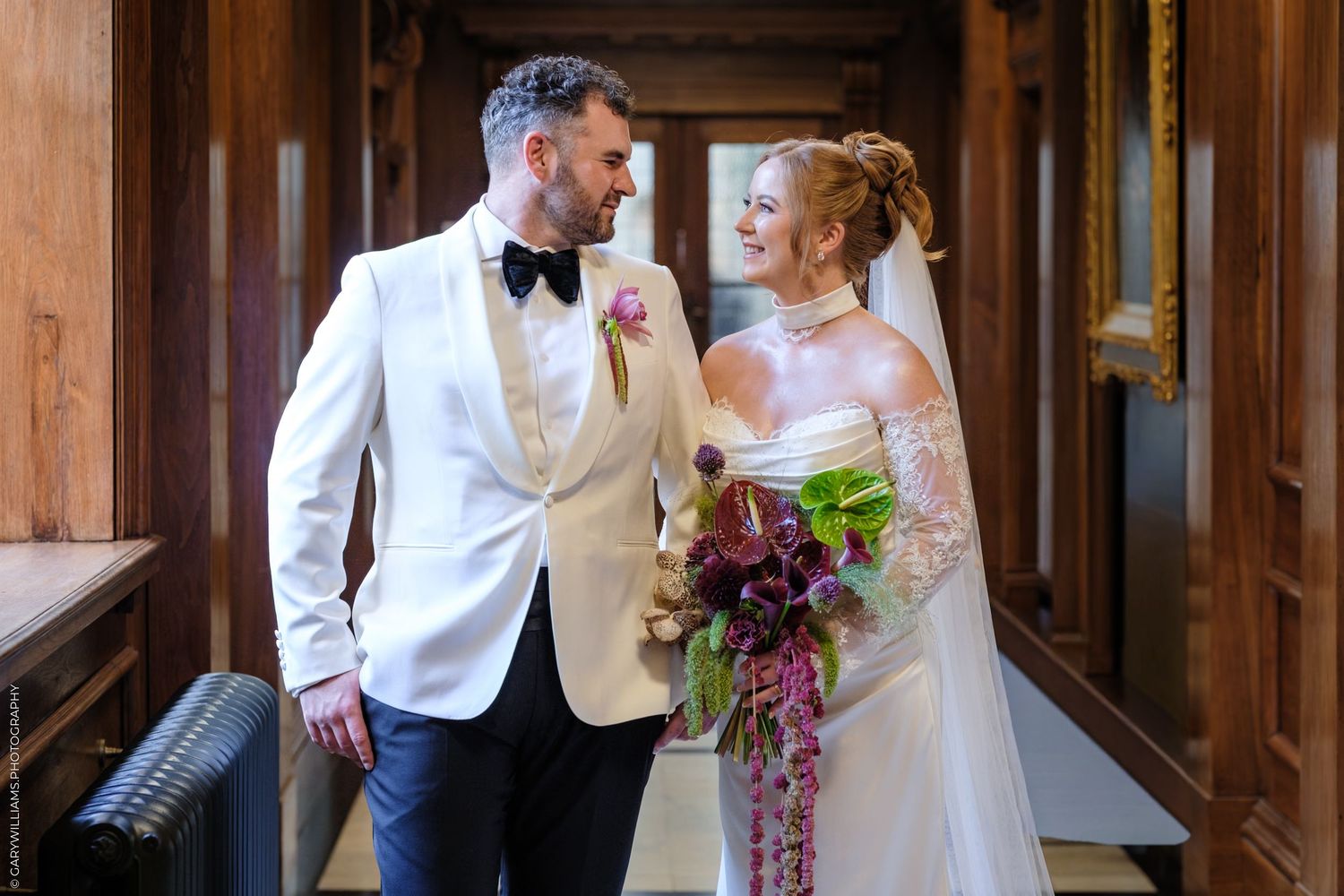 A couple in formal wedding attire share an intimate moment in a wooden paneled hall.
