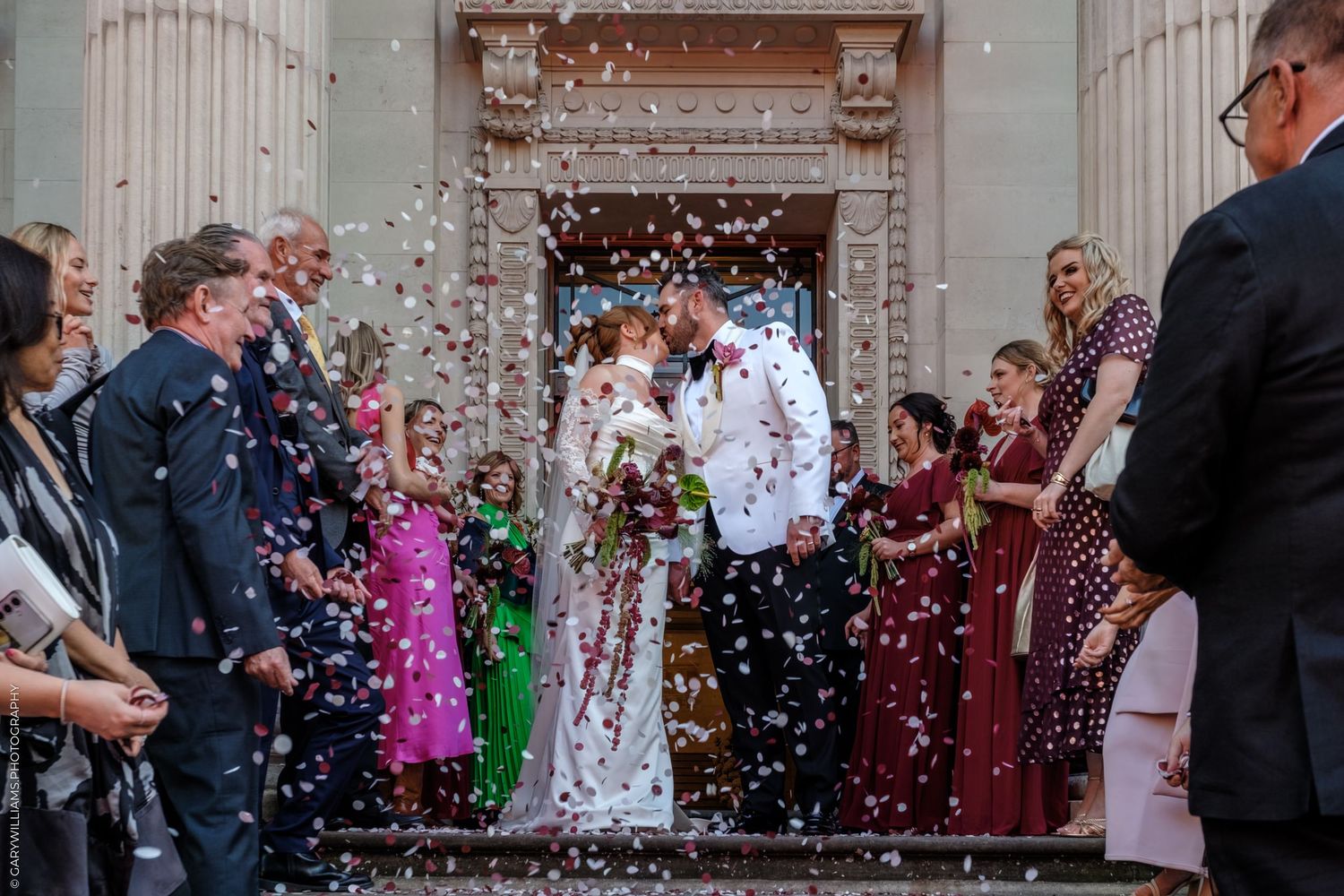 Wedding guests showering newlyweds with confetti outside a grand building with white columns during a joyful celebration.