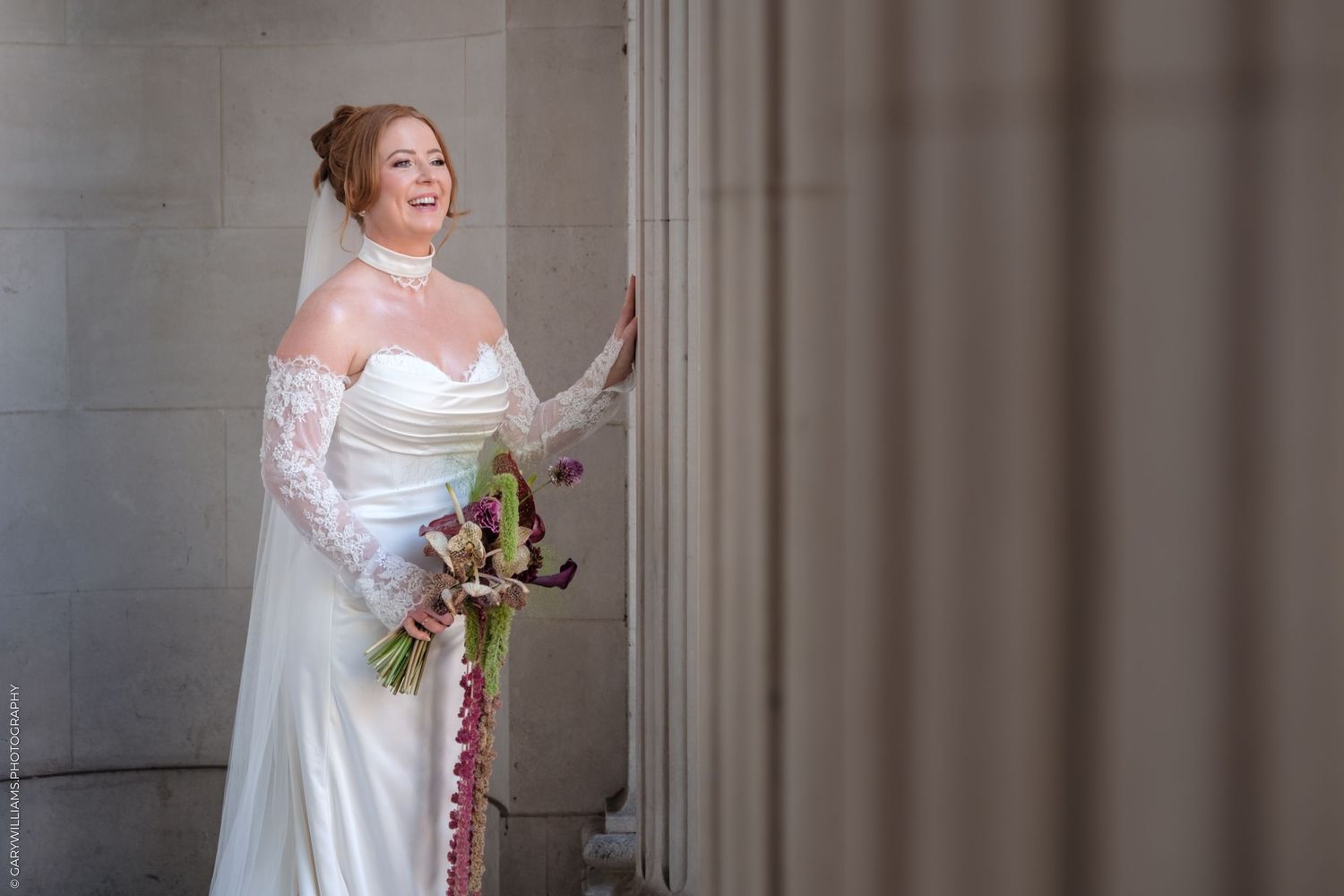 A person in an elegant white satin wedding dress poses in front of a stone wall interior.