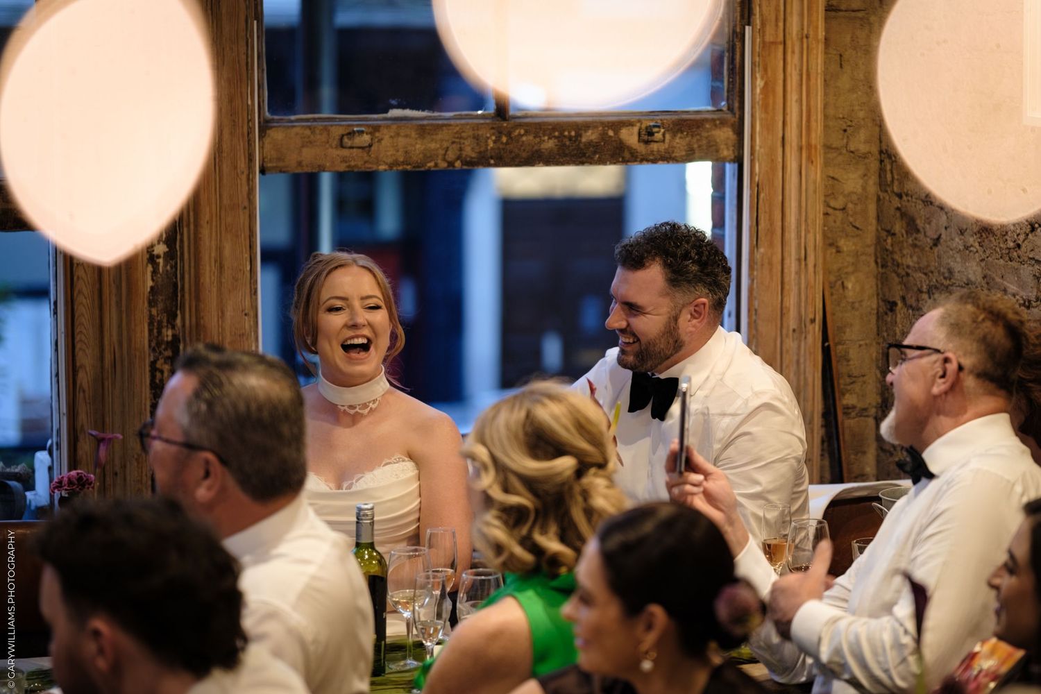 Wedding guests laugh and celebrate during a reception dinner with white paper lanterns hanging overhead.