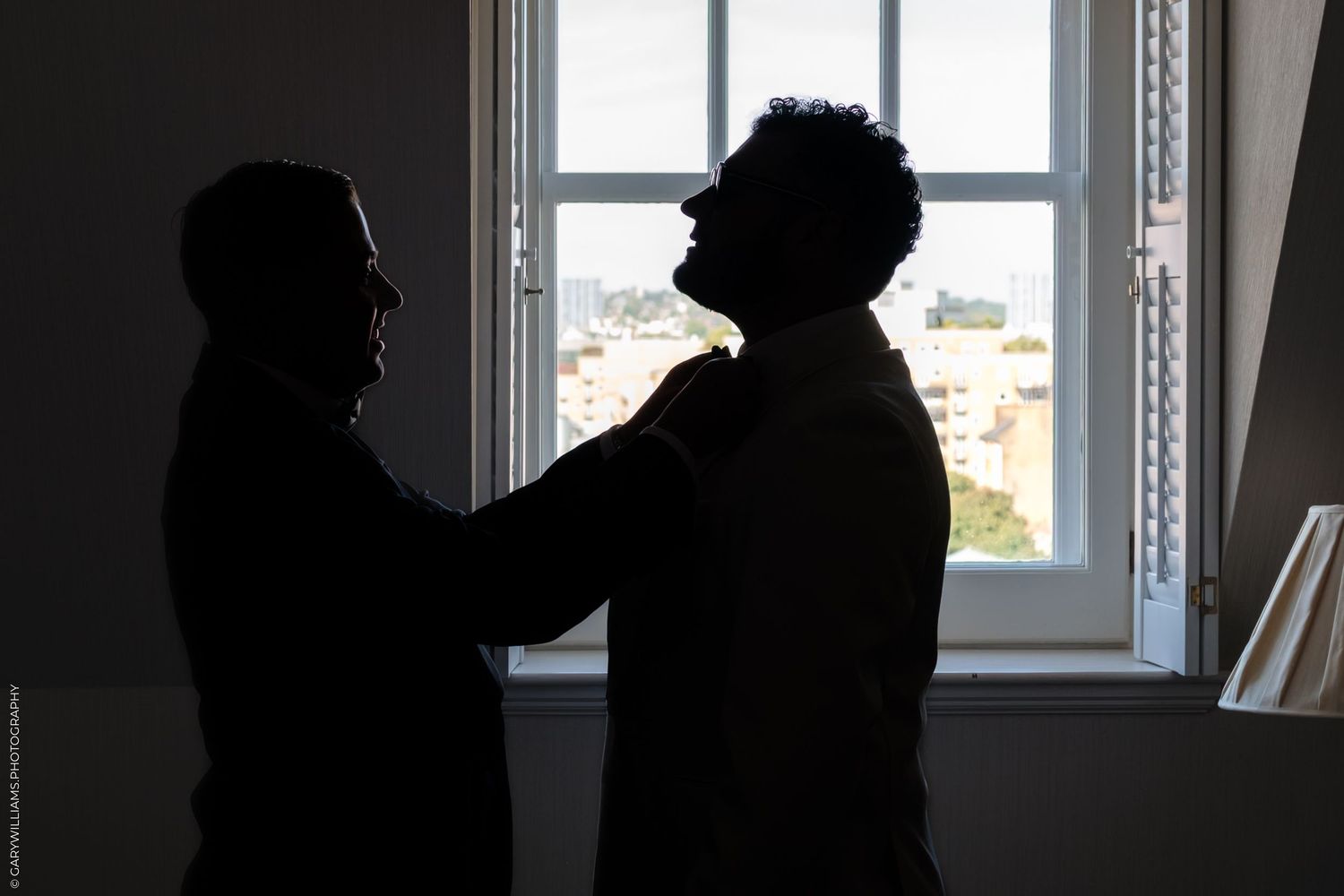 Silhouette of two figures standing by a window, one person appearing to adjust the other person's collar in dramatic lighting.