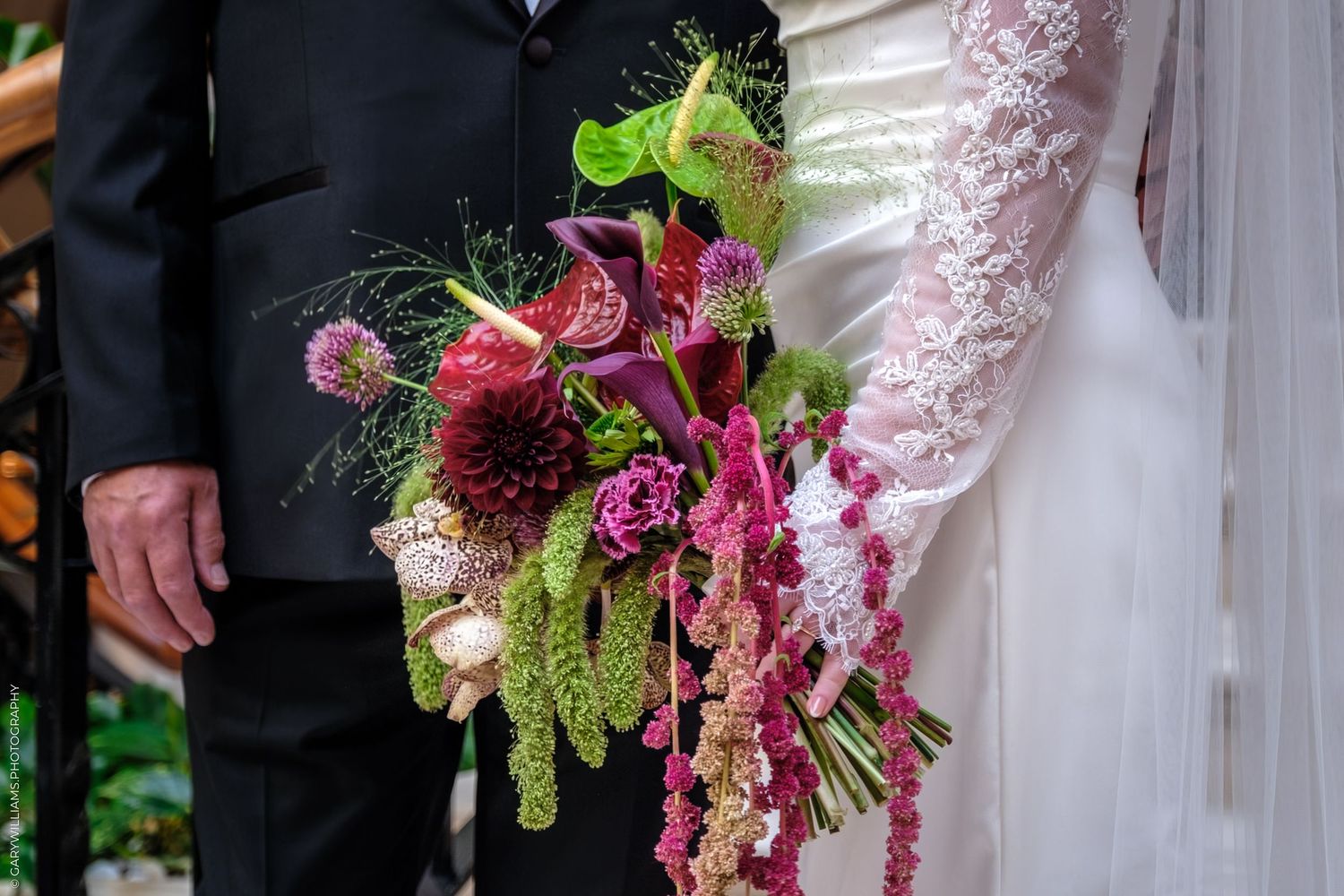 Wedding couple stands together with a vibrant cascading burgundy and pink floral bouquet against an elegant backdrop.