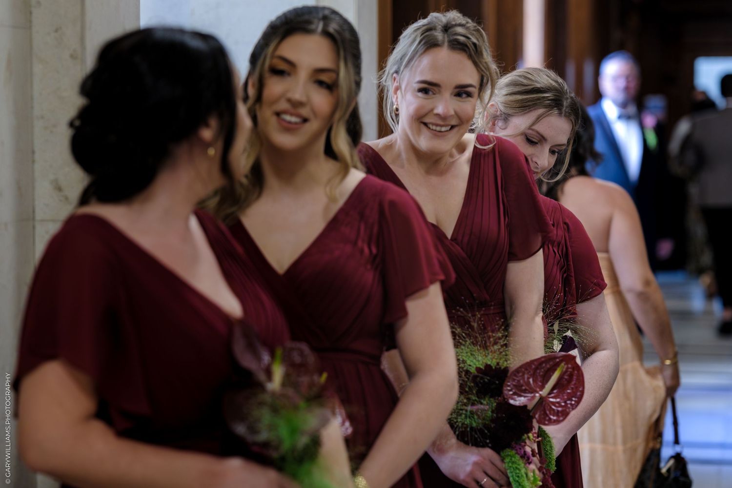 A row of people in burgundy dresses stand together smiling and holding small floral bouquets.