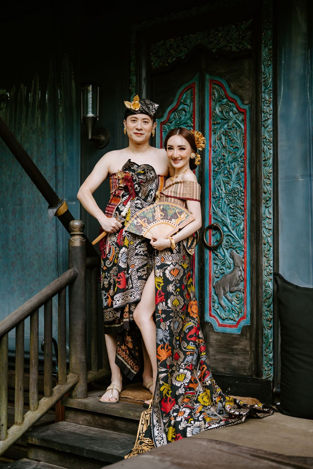 Two people pose in colorful patterned dresses on wooden porch with ornate teal walls.