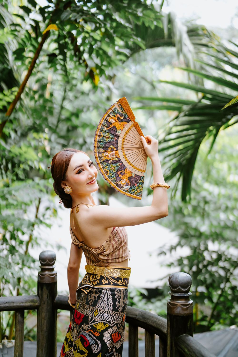 Person in traditional batik dress poses with decorative fan on wooden balcony.