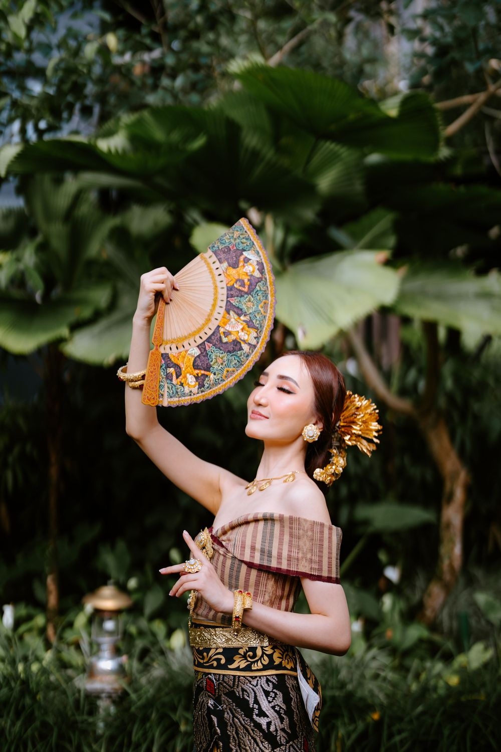 Person in ornate traditional dress poses dramatically with fan among tropical foliage.