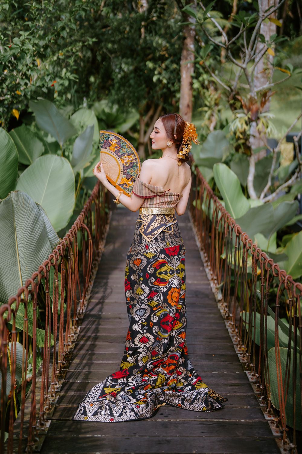 A vibrant floral dress stands out on a rustic wooden bridge surrounded by tropical foliage.