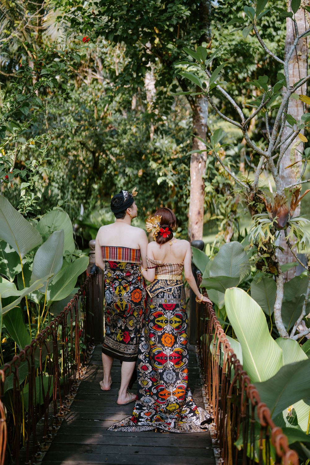 Two people walk along a wooden pathway surrounded by lush tropical plants and greenery.