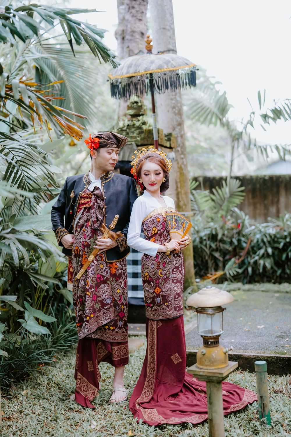 Couple in traditional Indonesian wedding attire posing in a tropical garden setting.