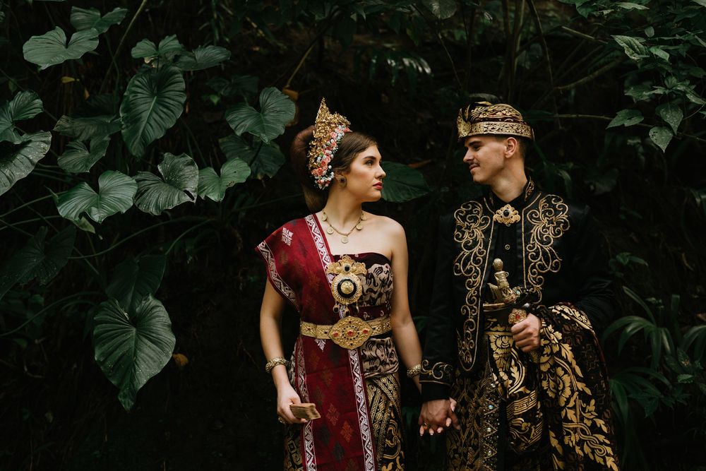 Couple in ornate gold and burgundy Balinese wedding attire pose against dark garden backdrop.