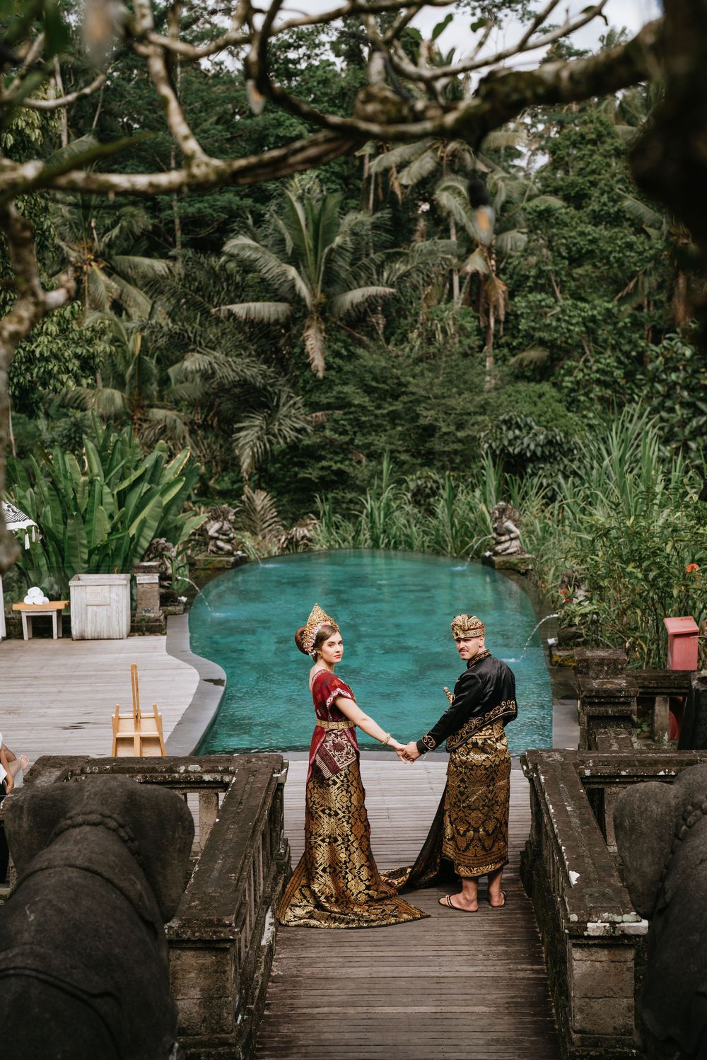 Couple in traditional Balinese wedding attire pose on wooden deck by turquoise pool surrounded by lush tropical garden.
