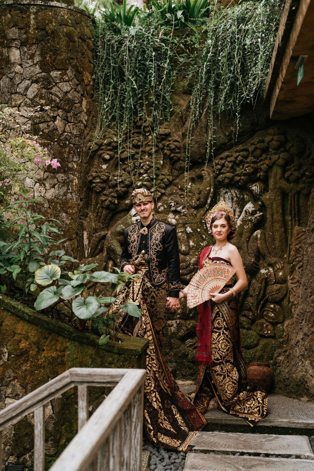 Traditional Balinese wedding photoshoot against stone wall backdrop with tropical foliage.