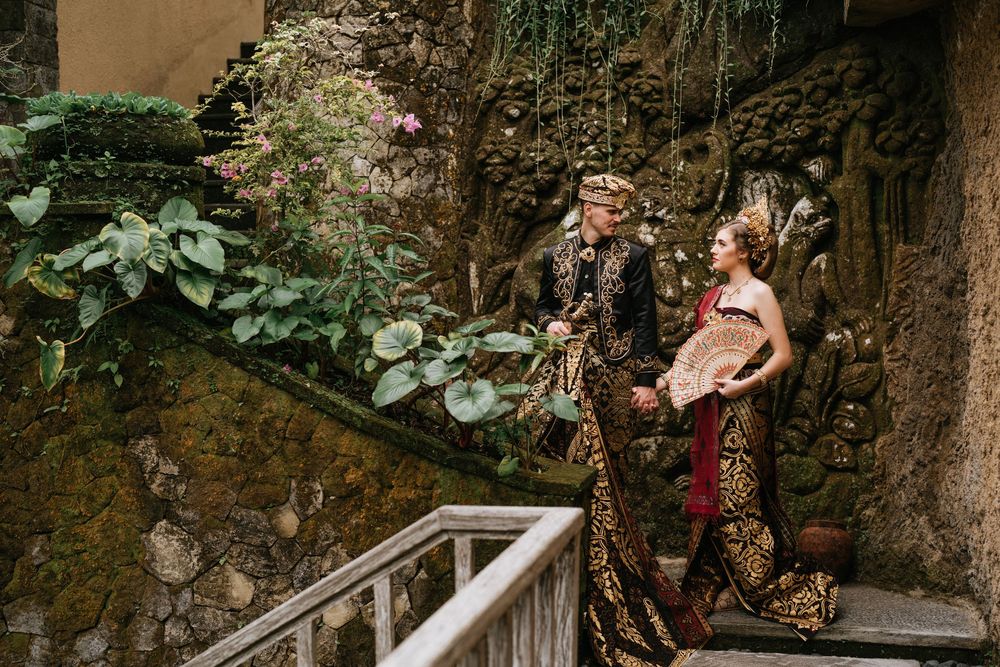 Wedding couple in ornate gold and burgundy traditional Balinese attire pose near stone walls.