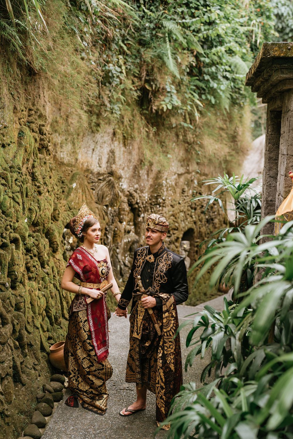 Couple wearing traditional Balinese wedding costumes walk along stone-walled garden path.