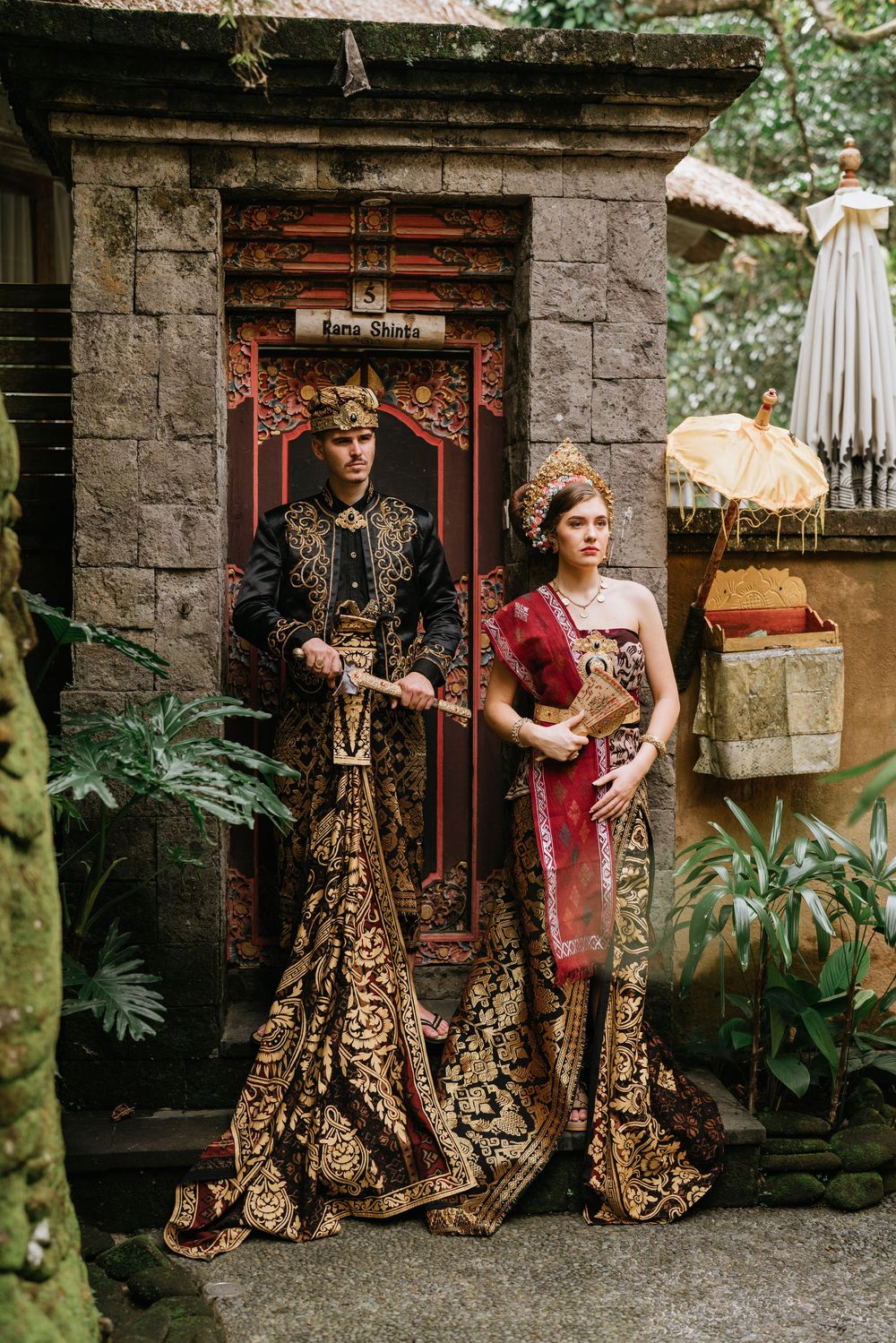 Wedding couple in ornate Balinese costumes pose in front of traditional stone temple entrance.