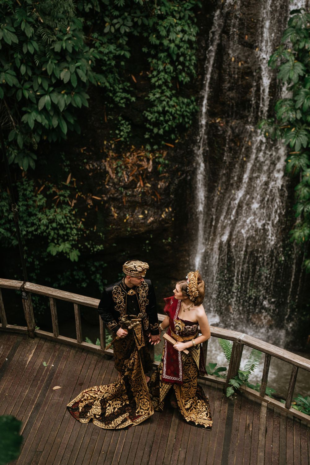 Couple in traditional Balinese wedding attire pose on wooden deck with waterfall backdrop.