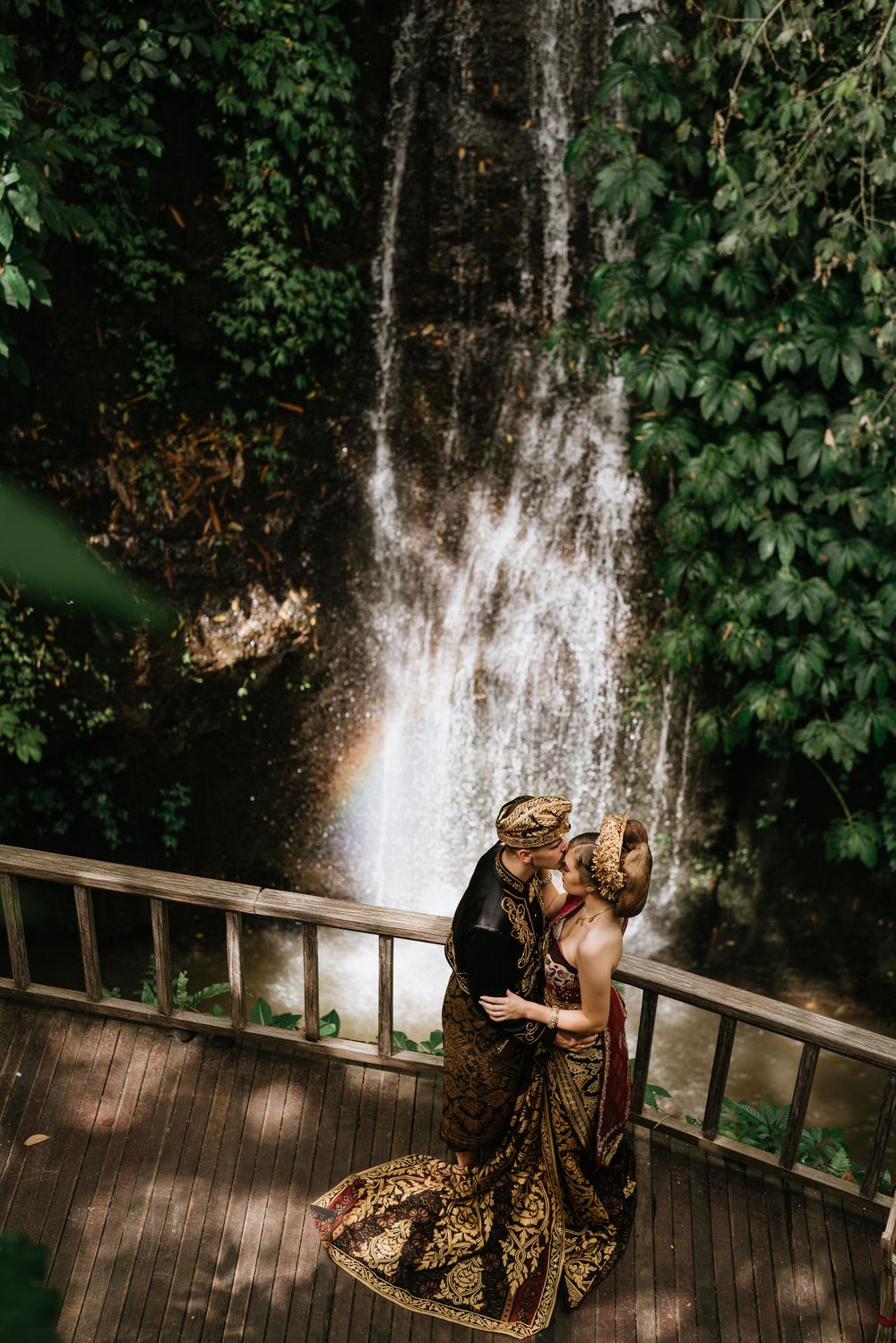 Romantic wedding photo on wooden deck with cascading waterfall and lush greenery.