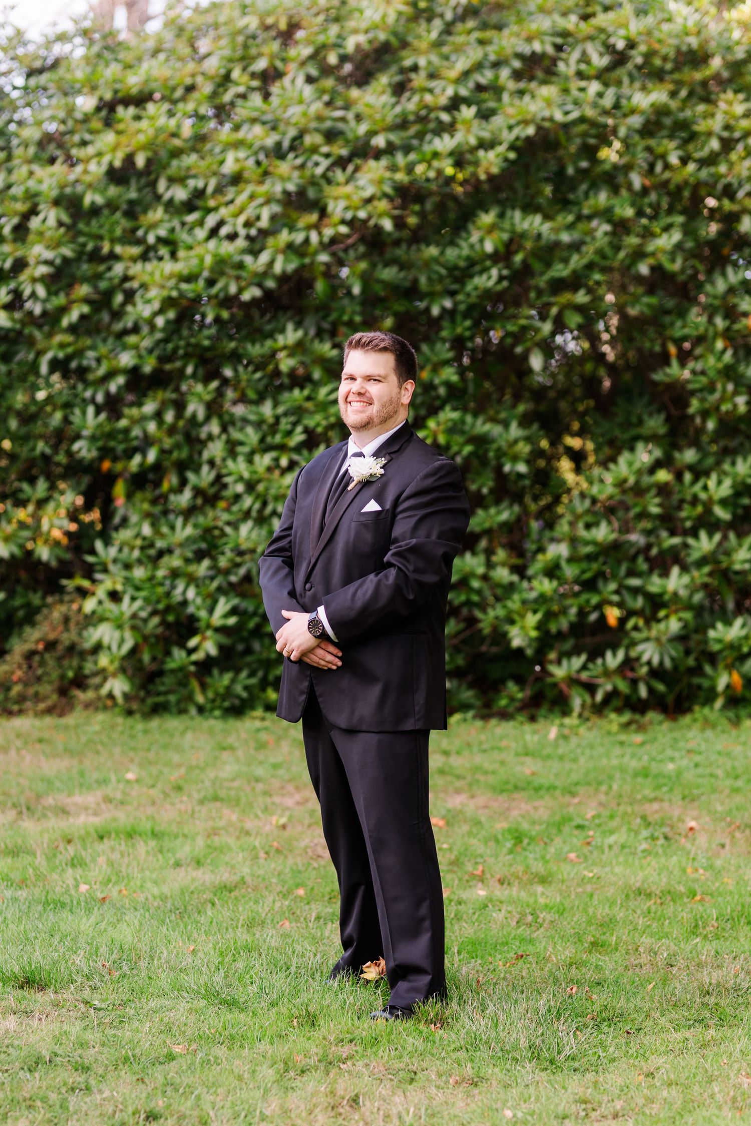 A groom in a black tuxedo poses outdoors against a leafy green backdrop during a wedding photo session.