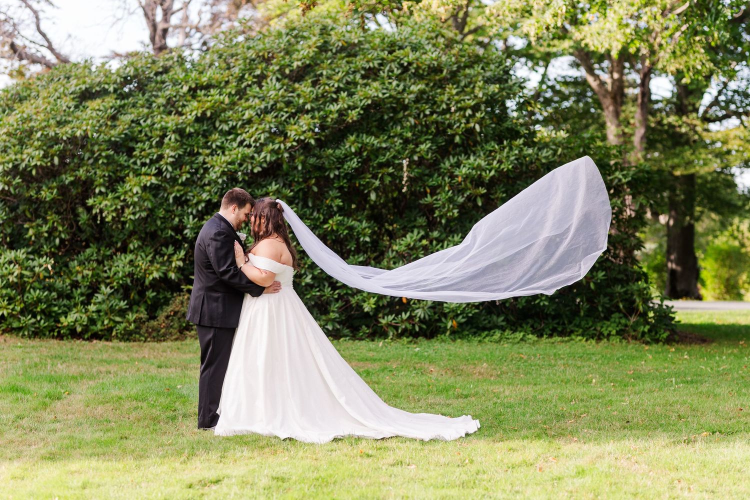 A couple in wedding attire stand on grass as a long white veil flows dramatically in the wind.