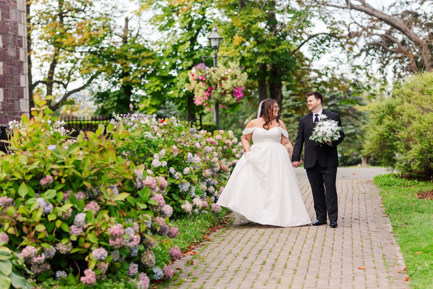 A couple walks along a garden path surrounded by blooming rose bushes at their outdoor wedding ceremony.