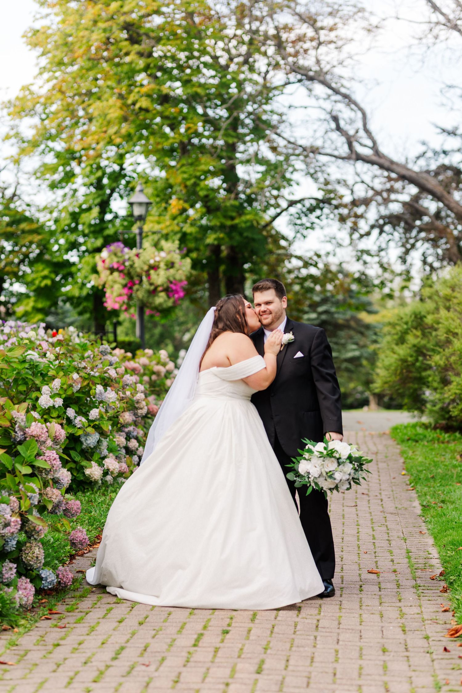 A romantic wedding couple poses on a brick pathway lined with blooming flowers and greenery.