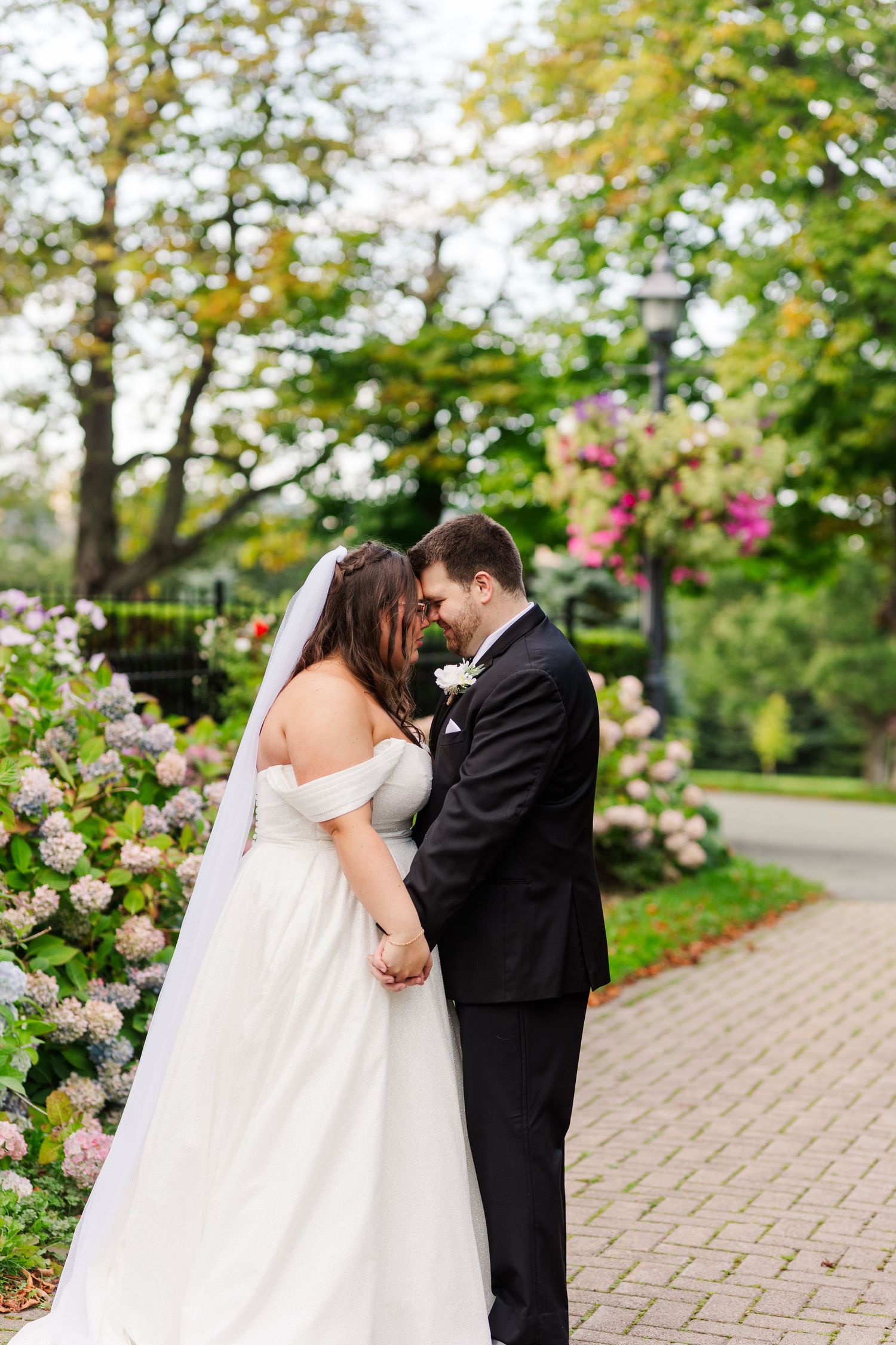 A romantic wedding couple shares a tender kiss among blooming pink roses in a garden pathway.