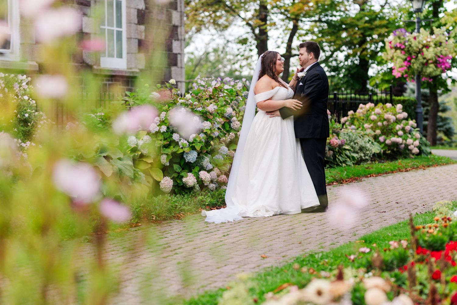 A couple shares a romantic moment on a garden path surrounded by blooming roses and flowers.
