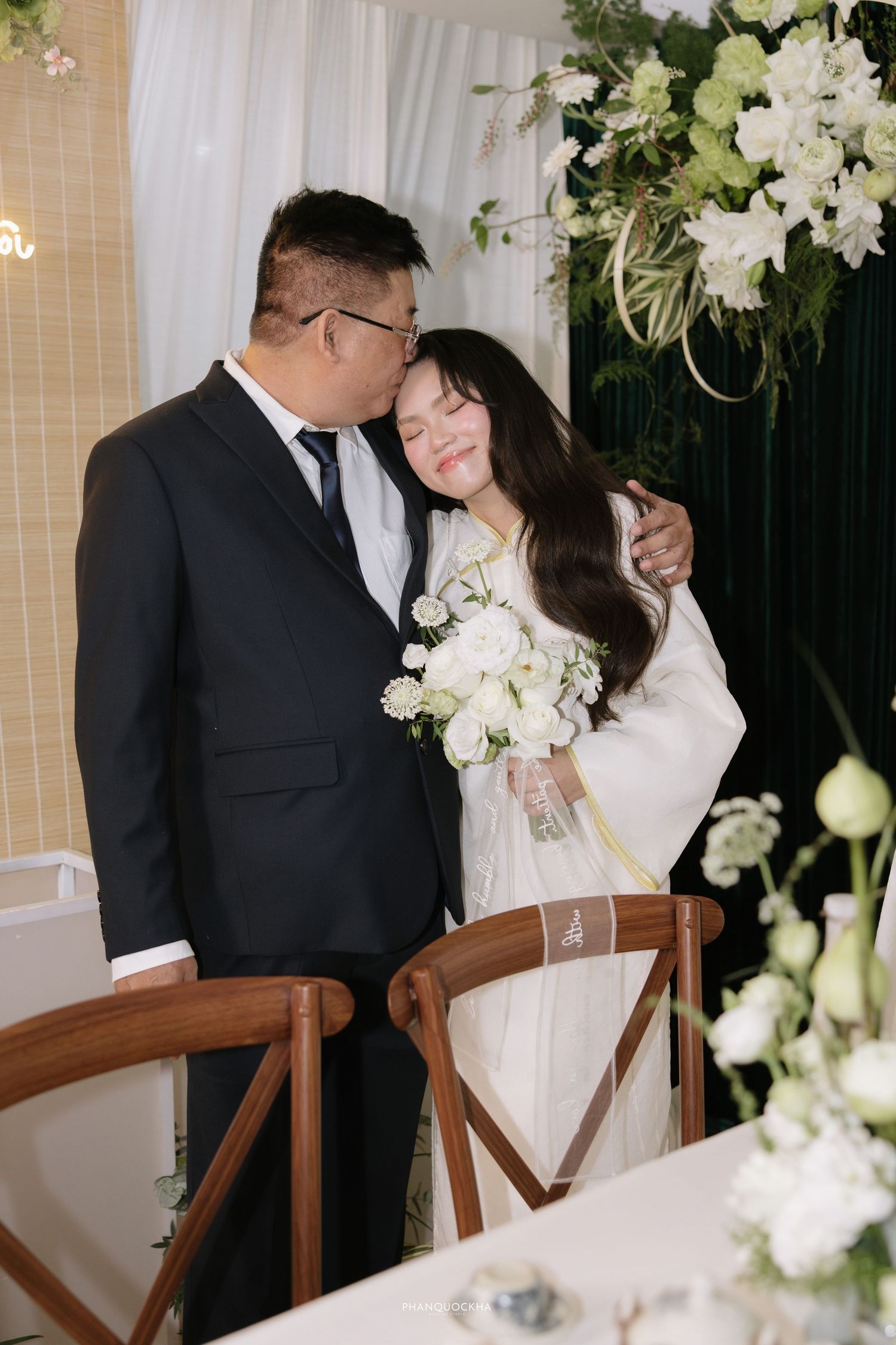 Groom in black suit embraces bride in white ao dai holding white bouquet at ceremony table.