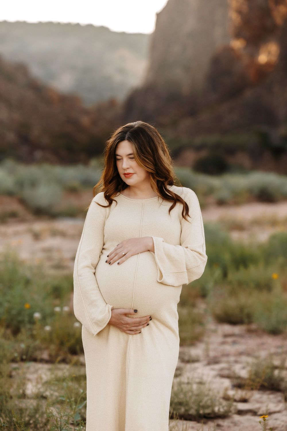 Pregnant woman in ivory dress cradling baby bump in scenic canyon setting.