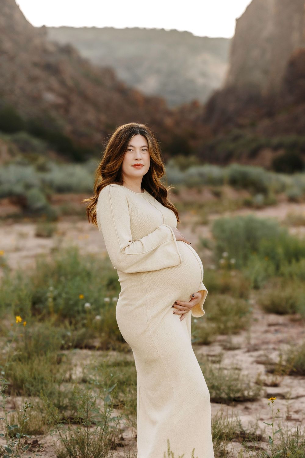 Elegant pregnant woman in cream fitted dress posing in desert canyon at sunset.