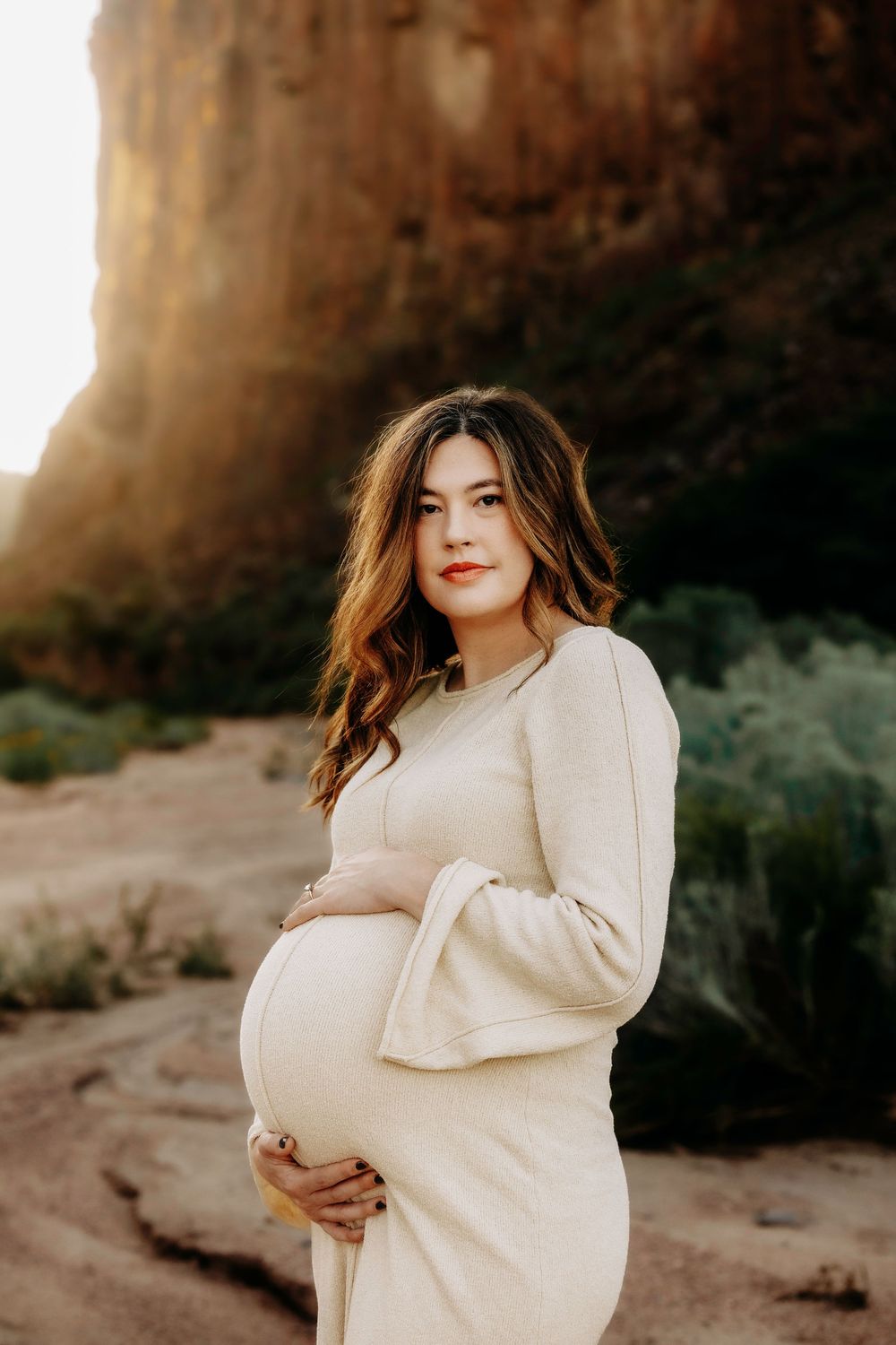 Pregnant woman in cream dress holding baby bump with golden canyon backdrop.