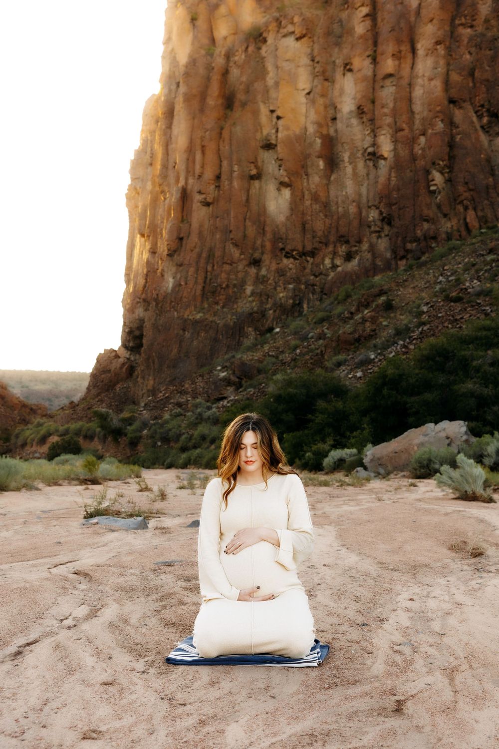 Pregnant woman in cream dress sitting peacefully on sandy ground near canyon wall.