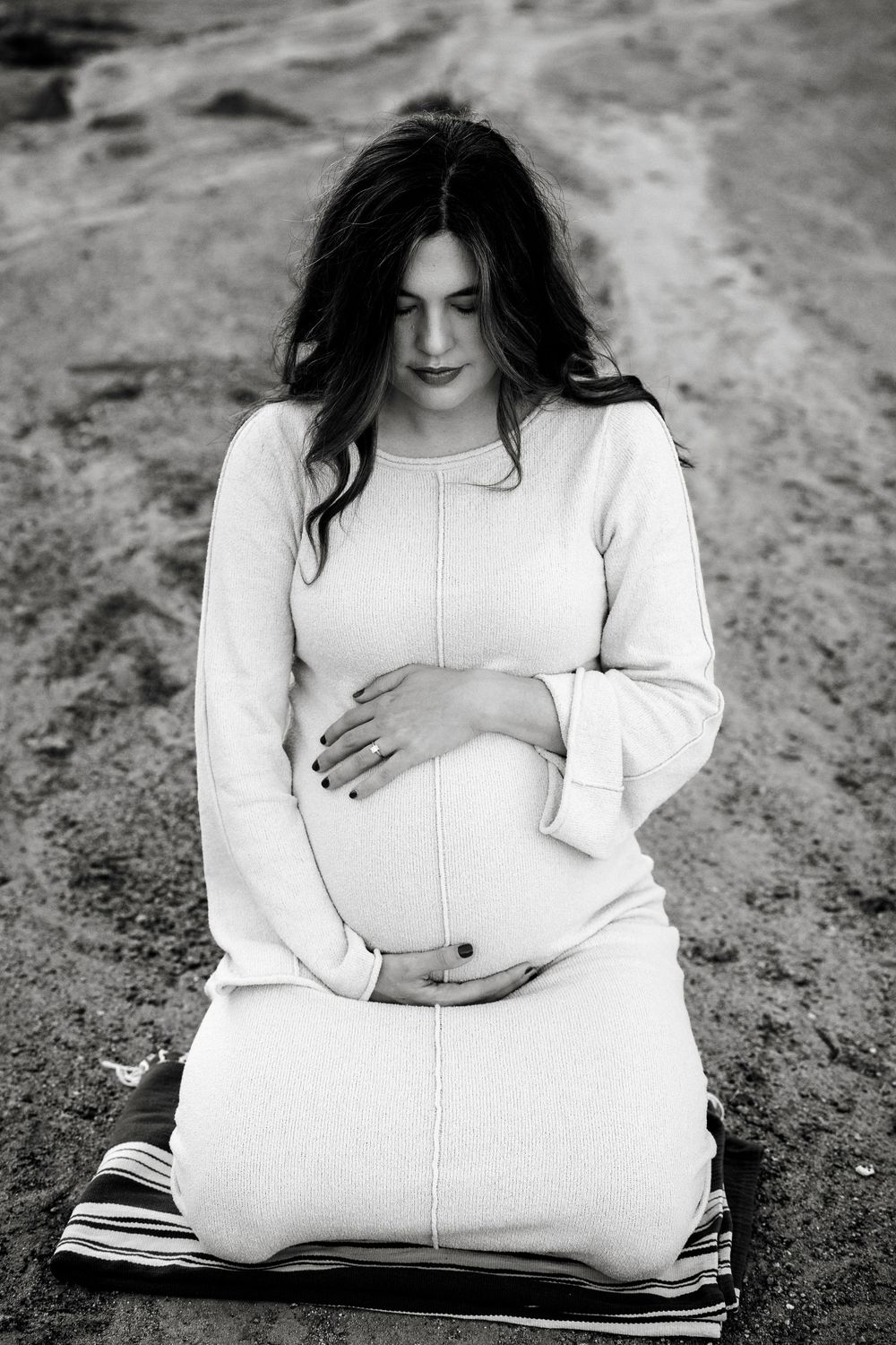 Black and white portrait of pregnant woman kneeling and cradling bump outdoors.