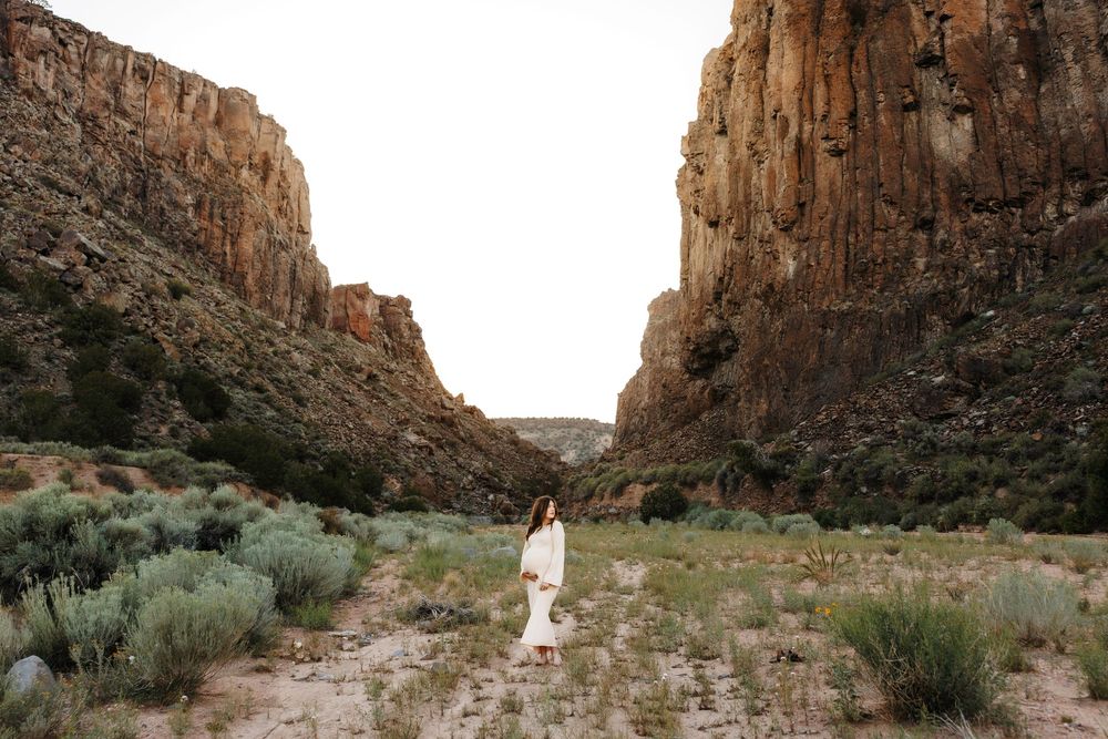 Pregnant woman in white dress standing between dramatic canyon walls surrounded by desert shrubs.