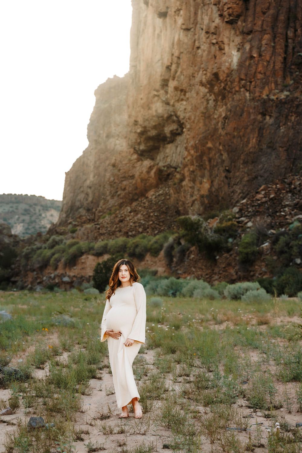 Glowing pregnant woman in cream dress holding belly near massive rocky cliff face at sunset.
