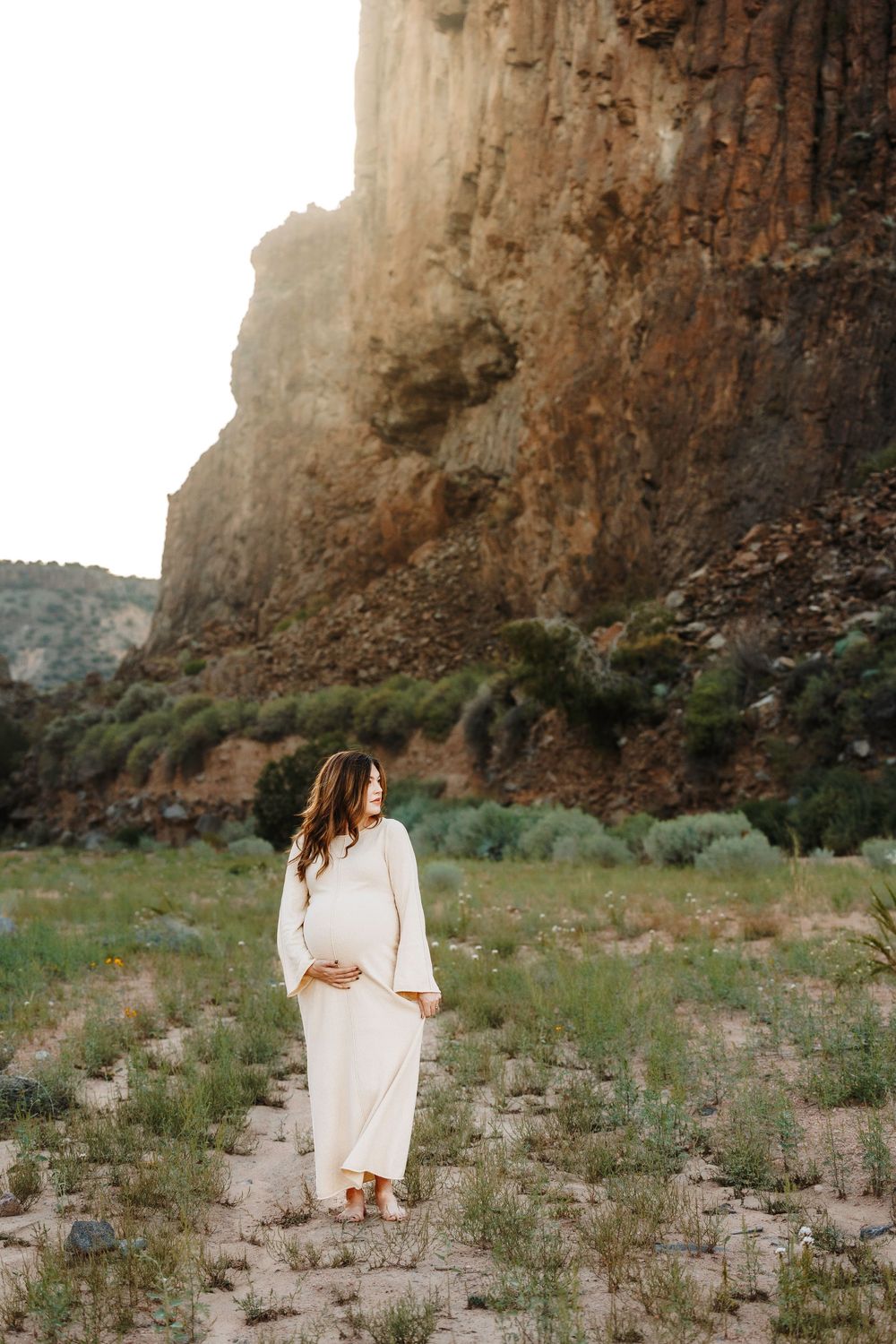 Pregnant woman in white dress cradling baby bump in scenic canyon with towering rock cliffs at golden hour.