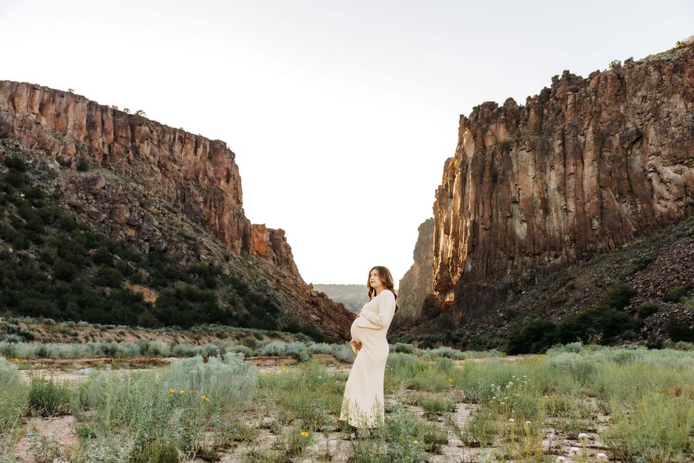 Pregnant woman in white dress standing between towering red rock canyon walls.