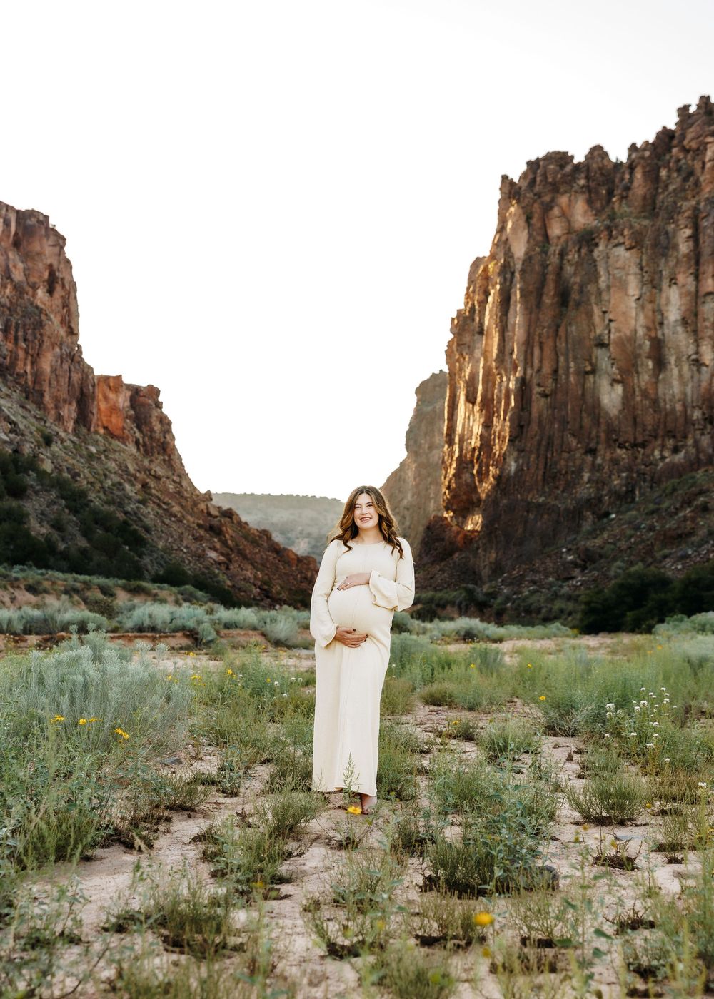 Smiling pregnant woman in white dress standing in a scenic desert canyon at golden hour.